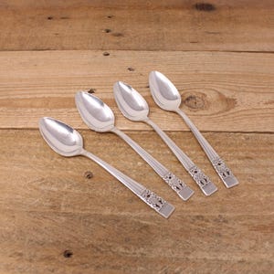 May include: Four silver spoons with decorative handles are displayed on a wooden surface. The spoons have a classic design with a rounded bowl and an ornate pattern near the handle's base. The spoons are arranged diagonally.