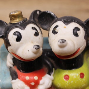 May include: Two vintage Mickey Mouse figurines, one wearing a hat, with black ears, white faces, and red and yellow clothing.