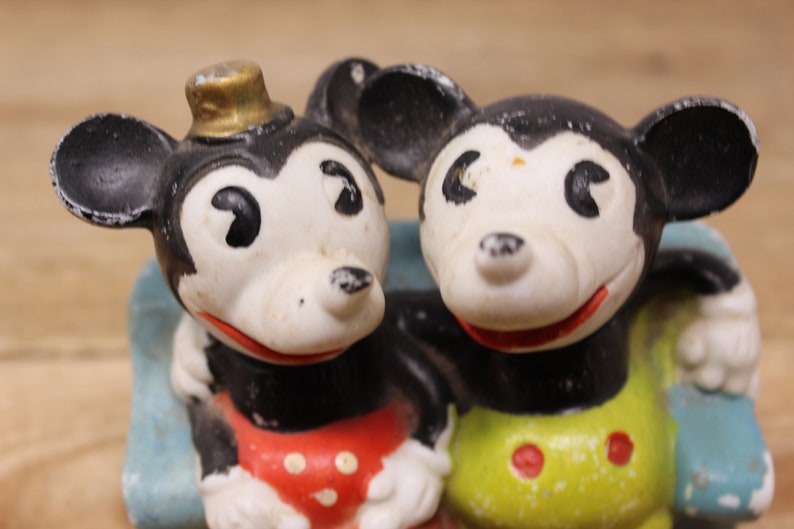 May include: Two vintage Mickey Mouse figurines, one wearing a hat, with black ears, white faces, and red and yellow clothing.