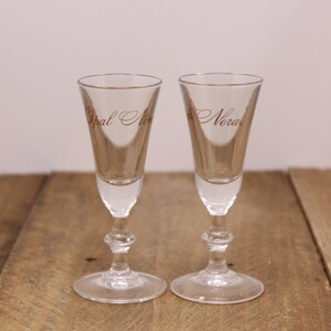 May include: Two clear, stemmed shot glasses with gold rims and text. The glasses have a bulbous base and a slender, conical bowl. The text on the glasses reads "Opal Nera". They are set on a rustic wooden surface.