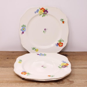 May include: Two cream-coloured ceramic plates with scalloped edges. Each plate features colourful floral designs in red, yellow, purple, and blue. The plates are displayed on a wooden surface.