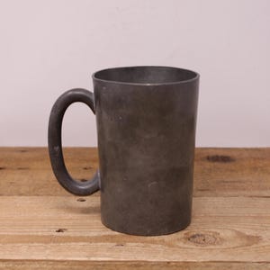 May include: A gray pewter mug with a rounded handle. The mug has a smooth, slightly textured surface.