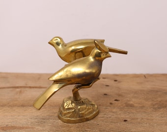May include: A pair of gold-toned brass bird figurines. The birds are perched on a small, leafy base. The birds are facing different directions, and one bird has its beak open.