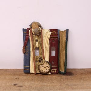 May include: Decorative light switch cover depicting a stack of books in blue, red, and green. A pocket watch and a small sculpture are attached to the books. The cover is designed to fit a standard light switch.
