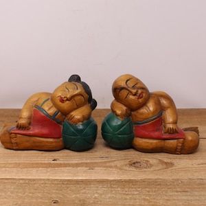 Peut inclure: Deux figurines en bois représentant des personnages endormis, chacun reposant sur une boule verte. Les figurines sont sculptées dans du bois de couleur claire, avec des accents rouges et noirs. Les figurines sont posées sur une surface en bois.