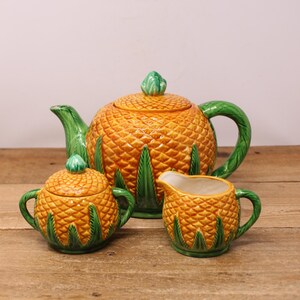 May include: A ceramic tea set comprising a teapot, sugar bowl, and creamer, each shaped like a pineapple. The set is predominantly orange with green detailing on the handles, lids, and leaves. The teapot features a lid with a green knob.