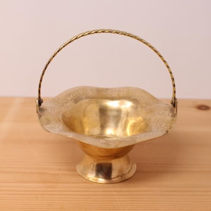 May include: A polished brass basket with a pedestal base and a twisted handle. The bowl features a scalloped edge and is decorated with an etched floral pattern around the rim. The metal has a warm, reflective gold tone throughout the entire piece.