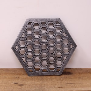 May include: A hexagonal, silver-colored trivet with a honeycomb pattern. The trivet is made of metal and has a distressed finish. It is designed to protect surfaces from hot items. The trivet is approximately 8 inches in diameter.