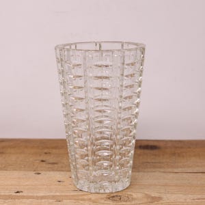 Peut inclure: Un vase en verre transparent avec un motif géométrique texturé. Le vase a une forme évasée et repose sur une surface en bois. Le vase est vide et semble vintage.