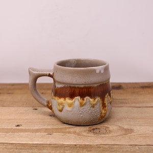 May include: A handcrafted ceramic mug with a handle. The mug features a light beige upper section, a band of brown in the middle, and a textured, dripped glaze in shades of brown and yellow. The mug sits on a wooden surface.