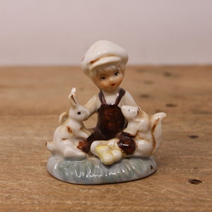 May include: A porcelain figurine of a boy wearing a brown overall and a white cap, sitting with two white rabbits on a blue and green base.