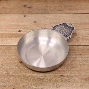 May include: A small, round, silver-coloured metal bowl with a decorative handle. The handle features a detailed relief design. The bowl sits on a wooden surface.