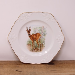 May include: White decorative plate with a scalloped edge and gold trim. The plate features a detailed illustration of a deer standing in a grassy area with trees in the background. The plate has embossed floral detailing.
