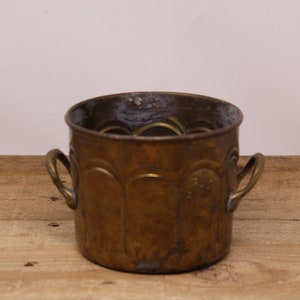 May include: A brass planter with two handles and a decorative embossed pattern. The planter is slightly tarnished and has a rustic look.