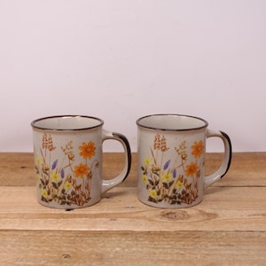 May include: Two ceramic mugs with a speckled beige finish and a brown rim and handle. Each mug features a floral design with orange, yellow, and purple flowers, along with brown and green foliage. The mugs are set on a wooden surface.