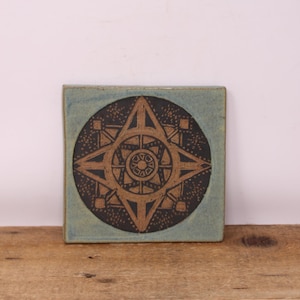 May include: Square ceramic tile featuring a geometric starburst design in brown and black. The tile has a light blue border and is displayed on a wooden surface. The starburst design is centered within a black circle.