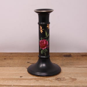 Peut inclure: Bougeoir noir avec un motif floral. Le bougeoir a une large base et un corps cylindrique. Une grande rose rouge et de plus petites fleurs jaunes sont peintes sur le corps. Le bord supérieur a un accent doré.
