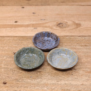 May include: Three small, handmade ceramic bowls in shades of blue and green. Each bowl has a unique, textured surface with embossed patterns. The bowls are arranged on a rustic, wooden surface, showcasing their artisanal quality.