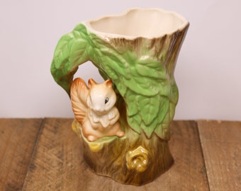 May include: A ceramic planter shaped like a tree stump with a squirrel peeking out from behind the leaves. The planter is painted in shades of brown, green, and white.