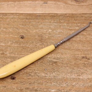 May include: An antique crochet hook with a cream-colored handle and a silver metal shaft. The hook is curved at the end. The tool is lying on a wooden surface.