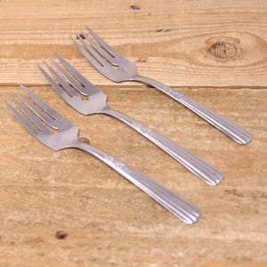 May include: Three stainless steel forks with a star-shaped cutout in the tines. The handles have a decorative pattern and are ribbed. The forks are arranged on a wooden surface.