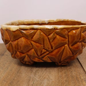 May include: A brown ceramic planter with a textured, geometric design. The planter has a rectangular shape with rounded corners and sits on three small feet. The rim is a lighter tan color.