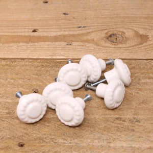 Peut inclure: Huit boutons de tiroir en céramique blanche avec un motif en relief décoratif. Les boutons ont une vis argentée au centre.