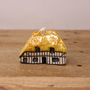 May include: A small ceramic cottage figurine with a thatched roof and white trim. The cottage has a black door and windows.