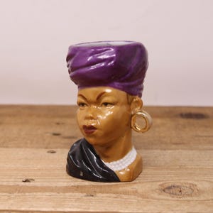 May include: A ceramic figurine of a woman's head, featuring a purple turban, gold hoop earring, and a black shawl. The face has brown skin, red lips, and a white pearl necklace. The figurine is on a wooden surface.