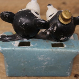 May include: A vintage ceramic toothbrush holder featuring two black and white cartoon dogs sitting on a blue base. The dog on the right is wearing a gold hat.