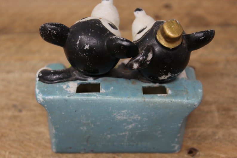 May include: A vintage ceramic toothbrush holder featuring two black and white cartoon dogs sitting on a blue base. The dog on the right is wearing a gold hat.