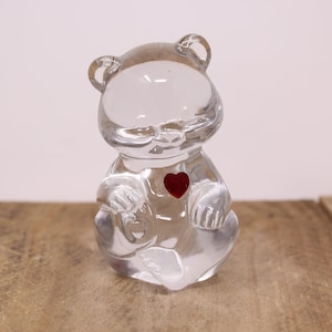 May include: A clear glass bear figurine with a red heart on its chest. The bear is sitting with its paws crossed.