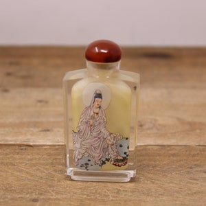 May include: A small, clear glass perfume bottle with a brown stopper. The bottle features a painted image of a seated figure in flowing robes, surrounded by a halo. The figure is depicted in soft colours, with a light yellow background.