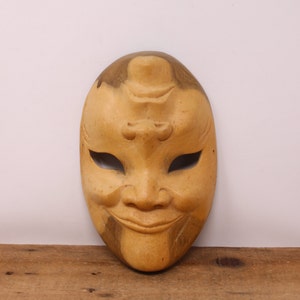 May include: A wooden mask with a carved face, painted in a light brown colour. The mask has a wide smile and large eyes.