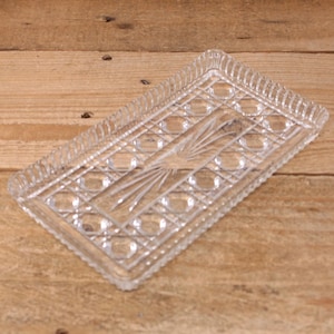 May include: A clear glass rectangular tray with a detailed geometric pattern. The tray has a central fan design and a decorative, ridged border. The glass is transparent, allowing the wooden surface beneath to be visible.