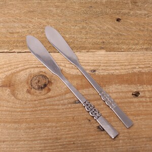 May include: Two stainless steel butter knives with rounded blades and decorative handles. The knives are placed diagonally on a wooden surface, highlighting their design. The handles have an ornate, swirling pattern.