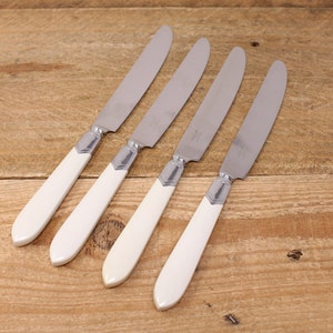 May include: Four butter knives with stainless steel blades and cream-coloured handles. The handles have a slight curve and are attached to the blades with a silver-coloured metal. The knives are arranged diagonally on a wooden surface.
