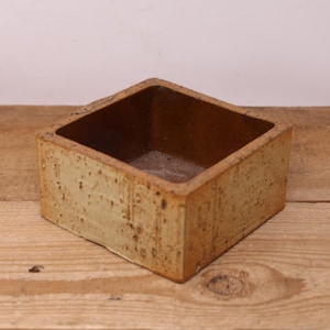 May include: A square ceramic planter with a light brown glaze and a speckled texture. The planter has a square opening at the top.