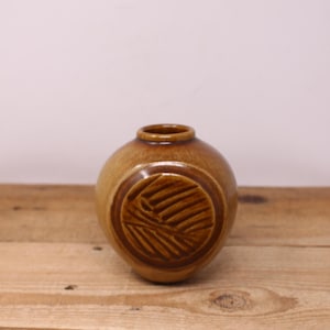 May include: A small, round, brown ceramic vase with a textured, circular design on the front. The vase has a narrow opening and sits on a wooden surface. The vase is a decorative item.