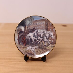 Peut inclure: Une assiette décorative ronde en céramique avec un bord doré, illustrant une pompe à incendie à vapeur tirée par des chevaux dans une rue. Un chien tacheté court au premier plan devant un bâtiment en briques représenté en arrière-plan.