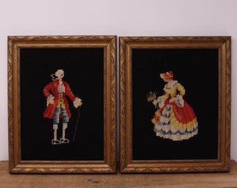 May include: Two framed needlepoint artworks. One depicts a man in a red coat and breeches with a cane, the other a woman in a yellow and red dress, holding flowers. Both are set against a black background within ornate gold frames.