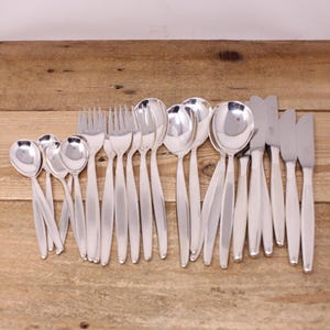May include: A selection of polished silver cutlery, including spoons, forks, and knives, displayed on a wooden surface. The silverware features a simple, elegant design with elongated handles and rounded spoon bowls. The knives have a slightly curved blade.
