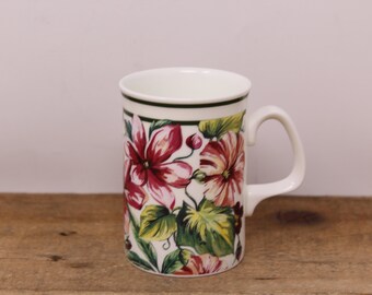 May include: White ceramic mug with a floral pattern of red and pink flowers, green leaves, and a dark green rim. The mug has a white handle and is on a wooden surface.