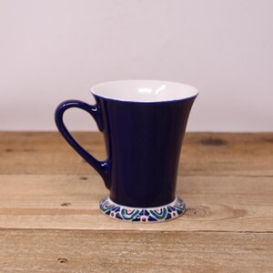 May include: A navy blue ceramic mug with a white interior and a decorative base. The mug has a curved handle and a flared shape. The base features a colourful pattern with blue, green, and red accents. The mug is set on a wooden surface.