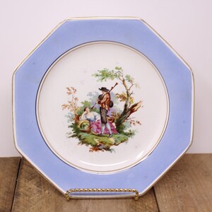 May include: An octagonal plate with a light blue border and a gold trim. The center of the plate features a pastoral scene with figures and trees. The plate is displayed on a gold-colored stand.