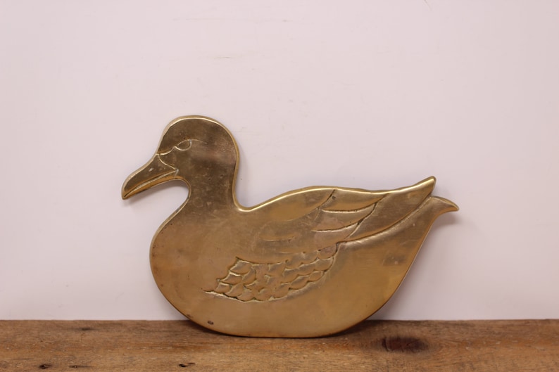 Pode incluir: Um descanso de panela de metal dourado em forma de pato. O pato tem um design detalhado com penas e uma asa.