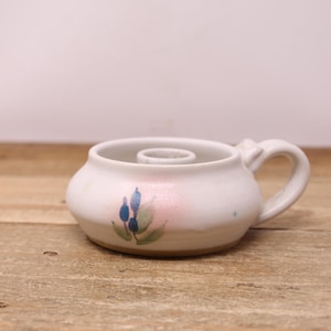Peut inclure: Un bougeoir en céramique blanche fait à la main, avec une petite poignée et un motif floral bleu et vert. Le bougeoir est posé sur une surface en bois.