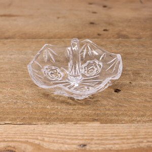 May include: A clear glass ring holder with a decorative design and a handle. The holder is sitting on a wooden surface.