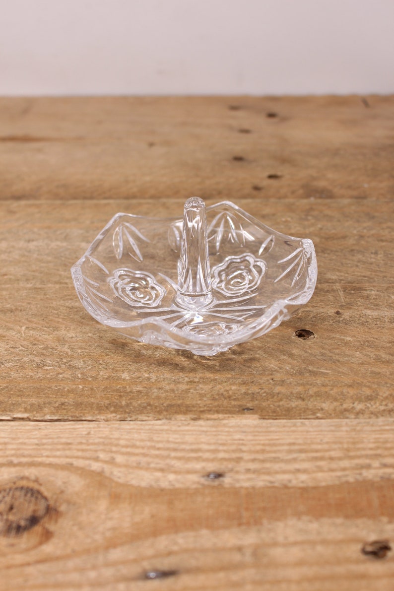 May include: A clear glass ring holder with a decorative design and a handle. The holder is sitting on a wooden surface.