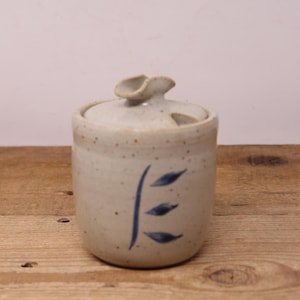 May include: A white ceramic sugar bowl with a blue and white leaf design. The bowl has a lid with a small handle.